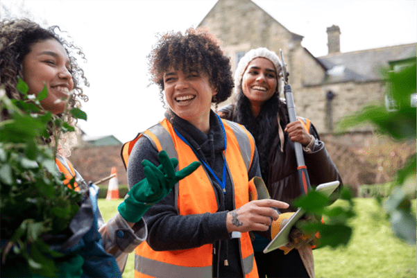 3 outdoor volunteers, stood in garden holding gardening tools, and also holding an electronic tablet