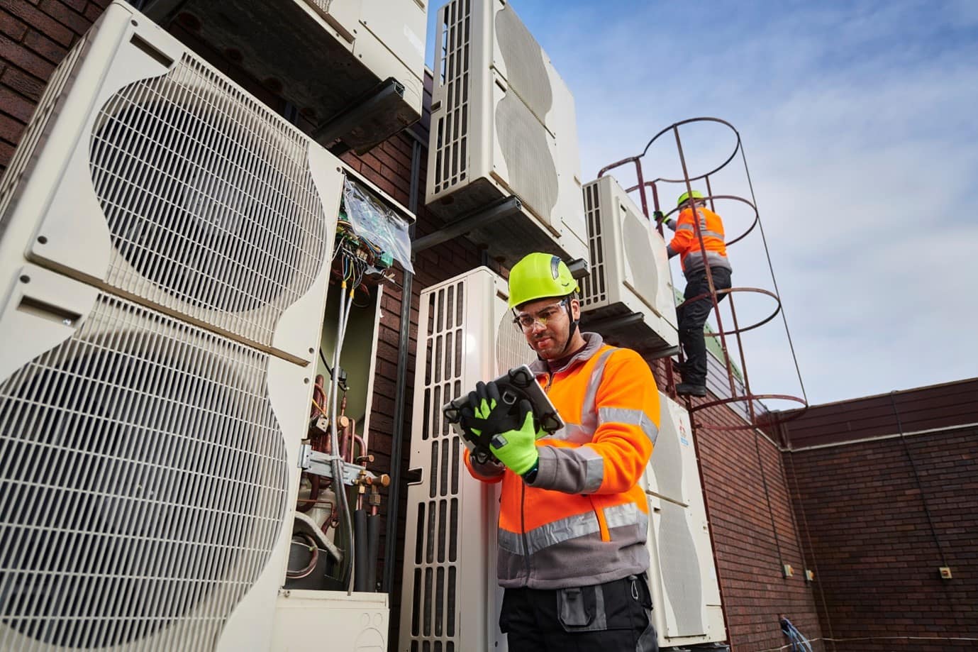 Two trades people, one stood next to an air ventilation system, one climing up some ladders behind