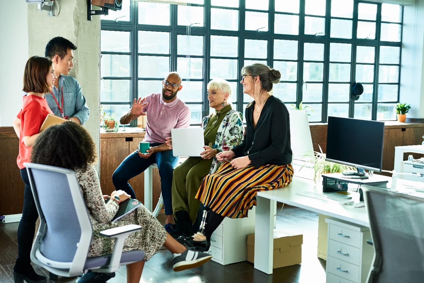 A group of people sat in an office having a discussion, a few people are sat on a white desk, a few people are stood up, and one person is sat in an office chair