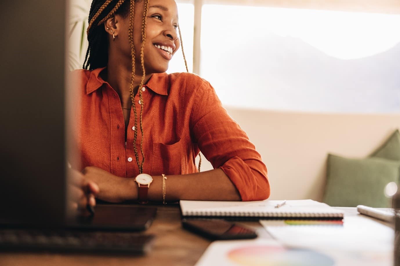Someone sat at a table smiling, with a notepad, computer and phone on the table