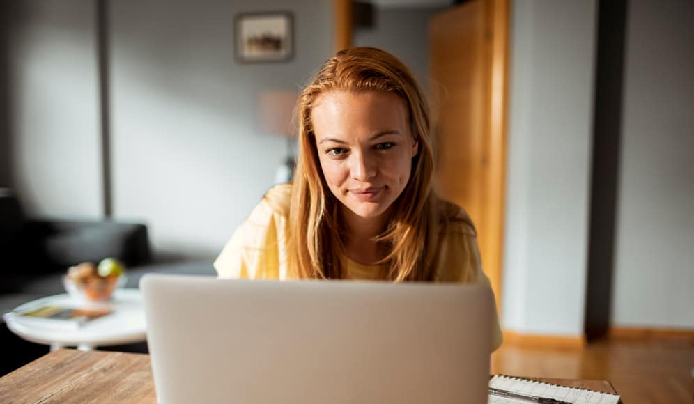 A person sat at a desk looking at a laptop screen, with a notebook and pen beside them