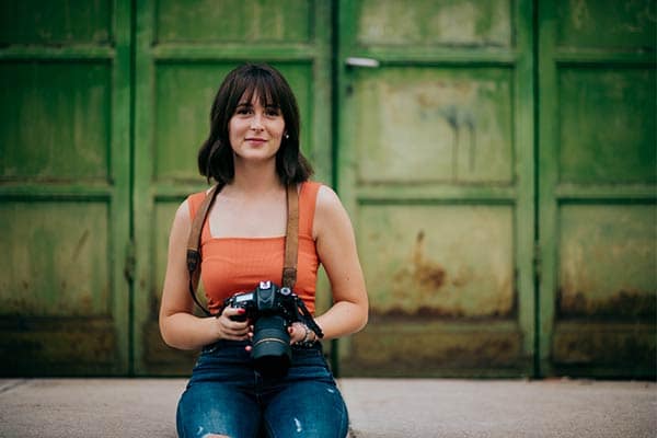 A freelance photographer wearing an orange top and smiling as she holds here camera.
