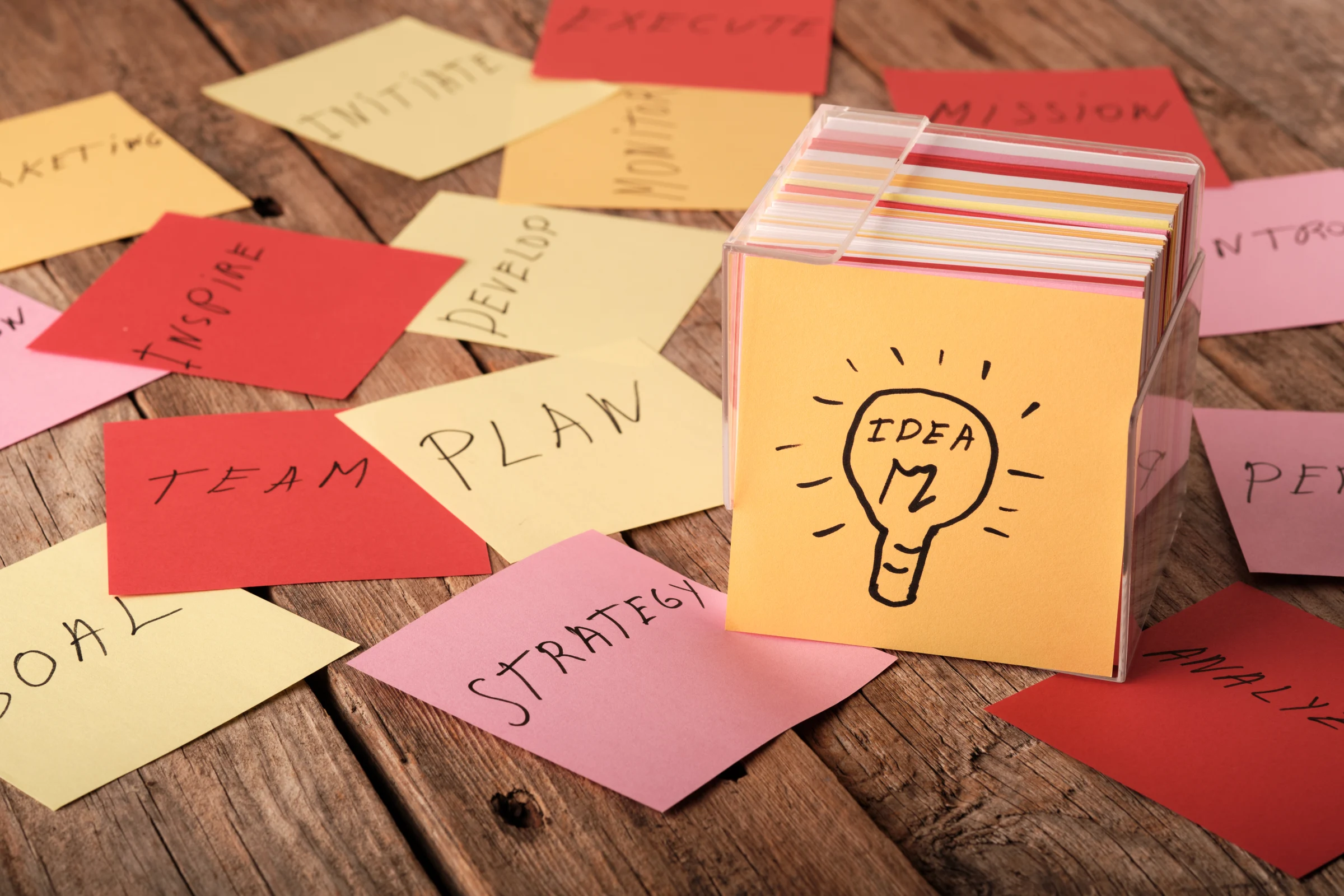 A box sits on a wooden surface filled with cards that include words about ideas, goals, strategy, and team planning. Various colored cards are scattered around the box.