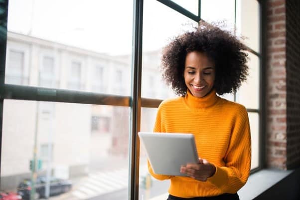 A lady stood infront of a window holding an electronic tablet and smiling.