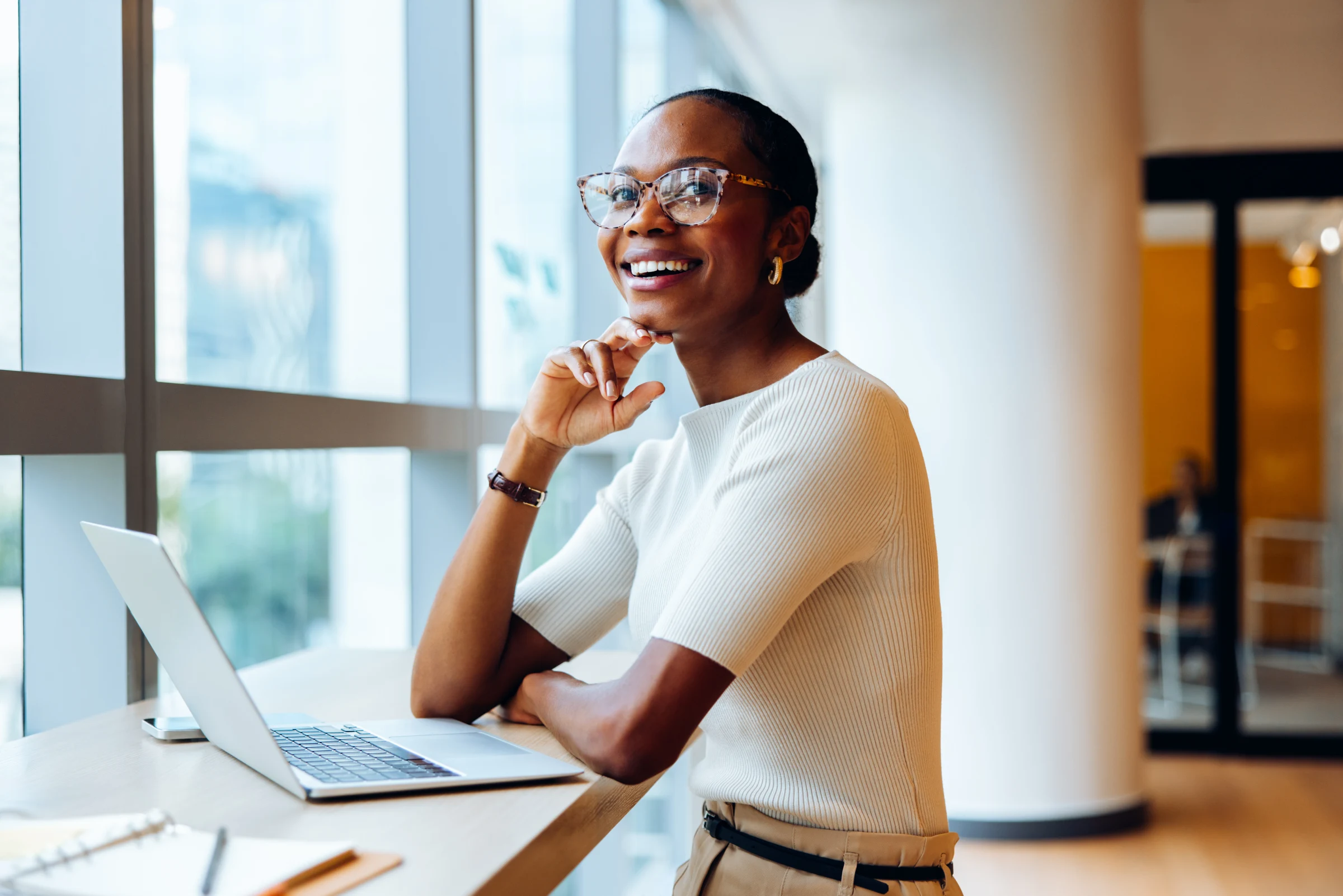 A cheerful young person wearing glasses sitting by a window with a laptop
