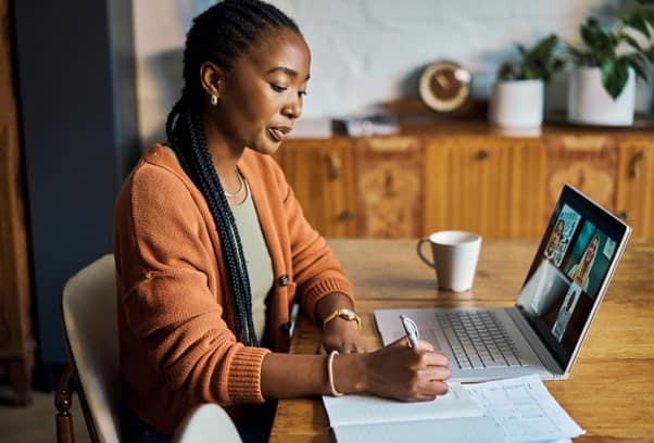 A lady sat at a table, writing in a notebook with her computer next to her
