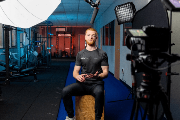 A man sat infront of cameras, wearing gym gear and talking, with a gym behind him