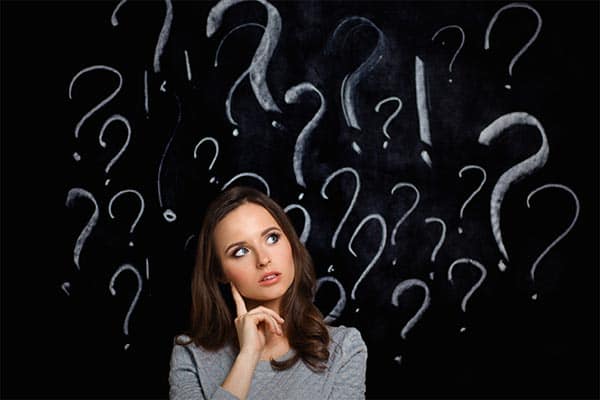 Someone looking inquisitive, stood in front of a blackboard with question marks 