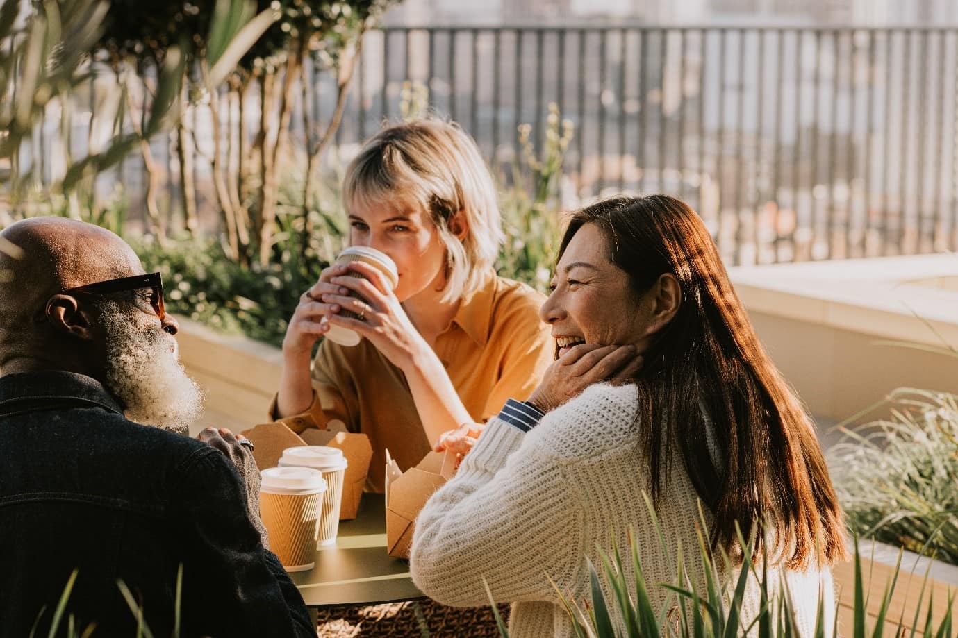 Three people sat outdoors smiling and talking whilst drinking coffee