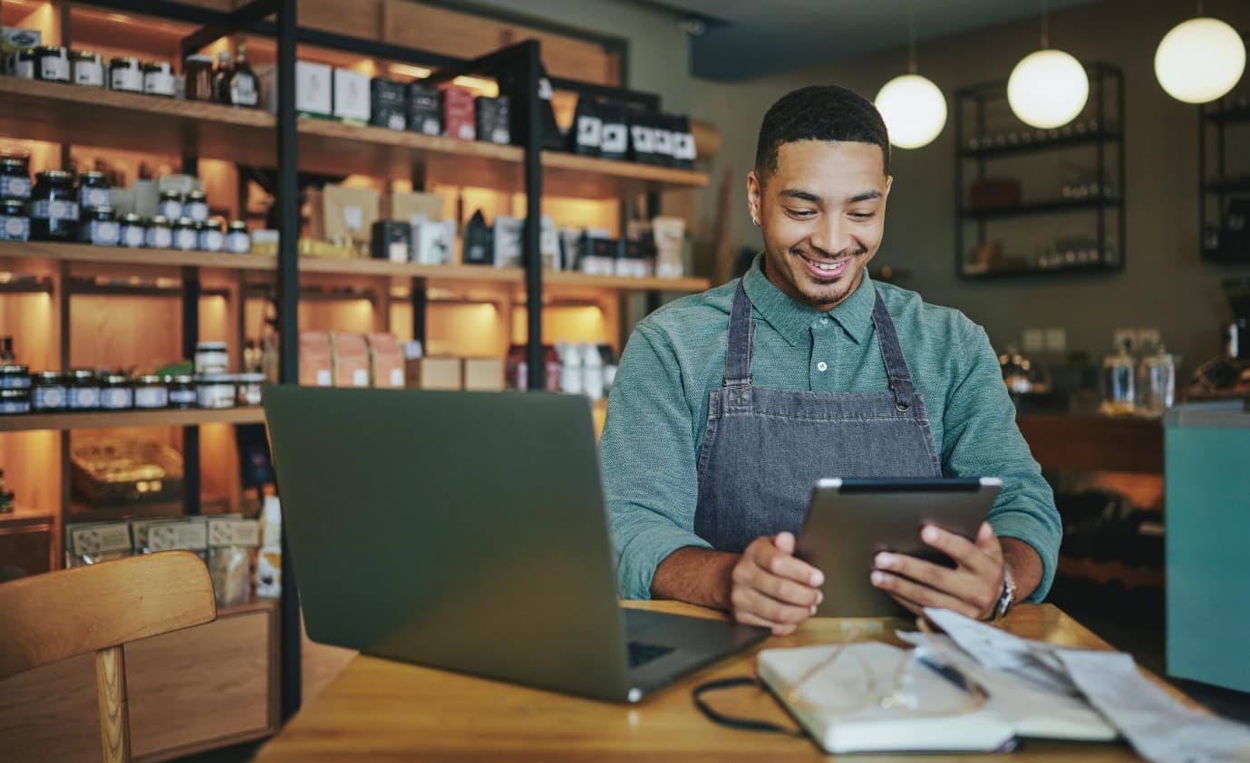 An employee at a coffee shop, holding an electric tablet next to a computer and some paperwork