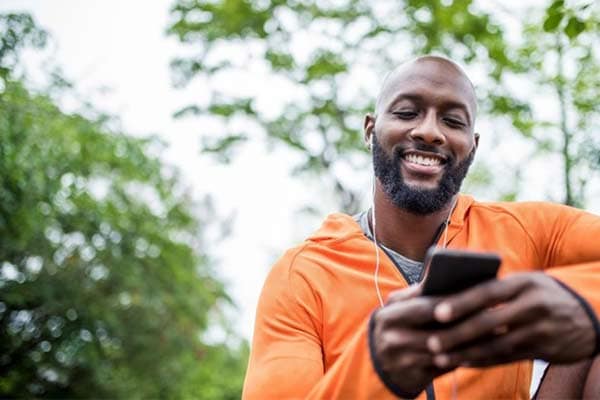 A man in an orange jacket listening to a podcast through his headphones while sat in a park.