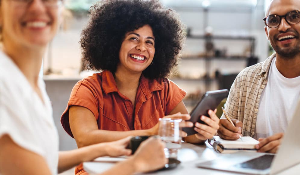 Three people sat at a table smiling, with one holding an electronic tablet