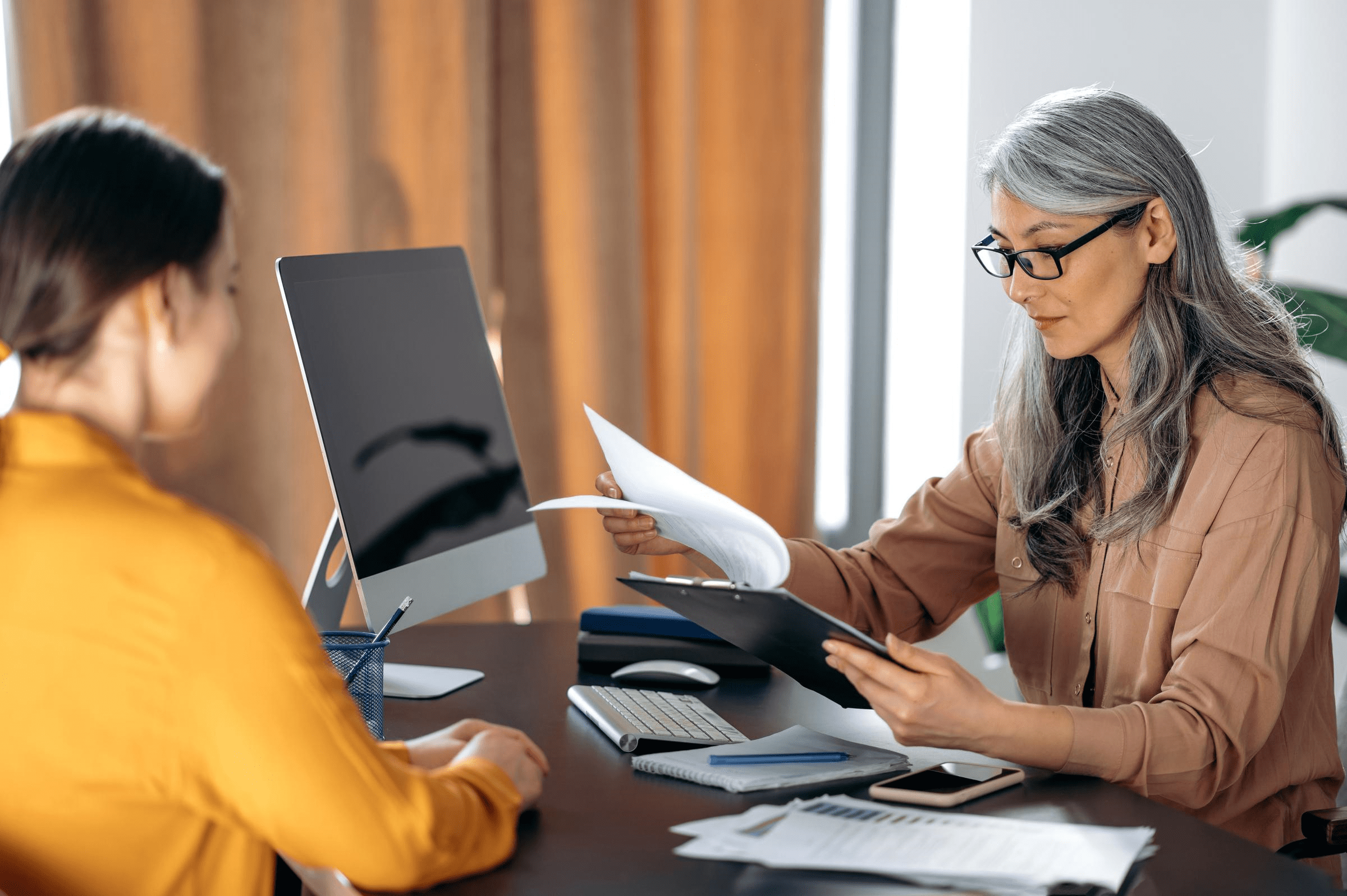 Two women looking at notes on a clipboard, whilst at a desk