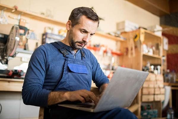 A male joinery business owner in overalls works on a laptop in his workshop.
