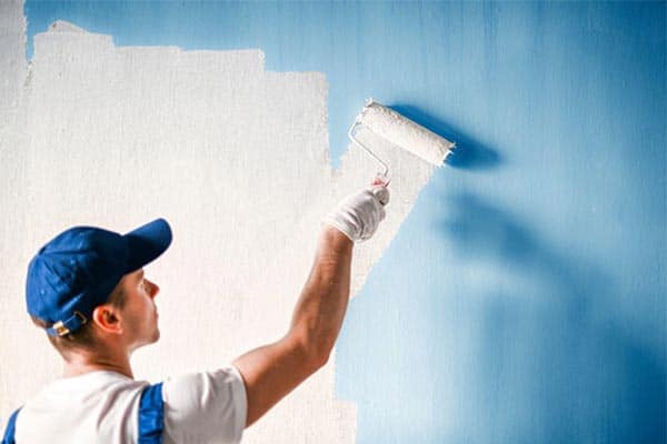 A painter and decorator uses a roller brush to paint a blue internal wall white.