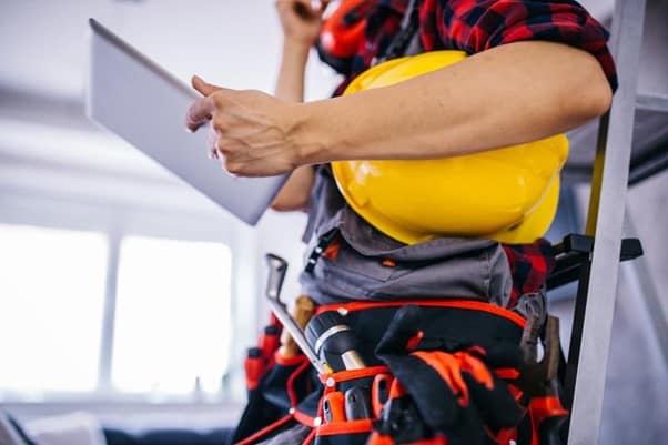 A tradesperson, with a tools belt and hard hat under his arm, holding an electronic tablet