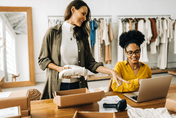 Two people infront of a rail of clothes, one is sat down facing a computer and the other is packaging an item of clothing