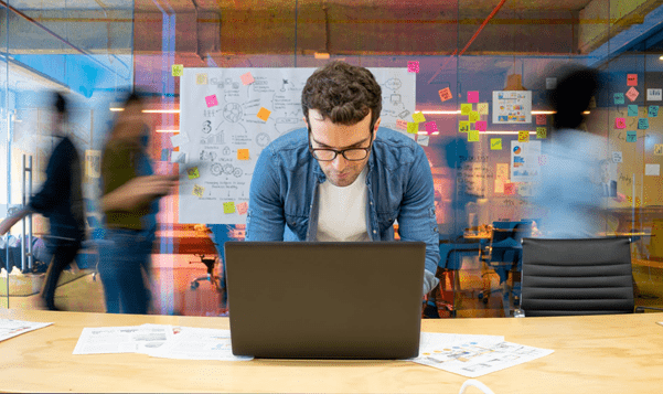 Someone stood infront of a laptop, with people walking in the background infront of a screen with a lot of post-it notes
