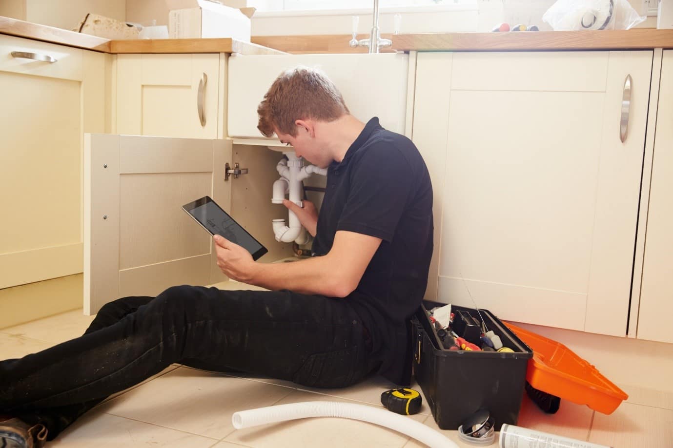 A plumber sat on a kitchen floor working on sink pipes