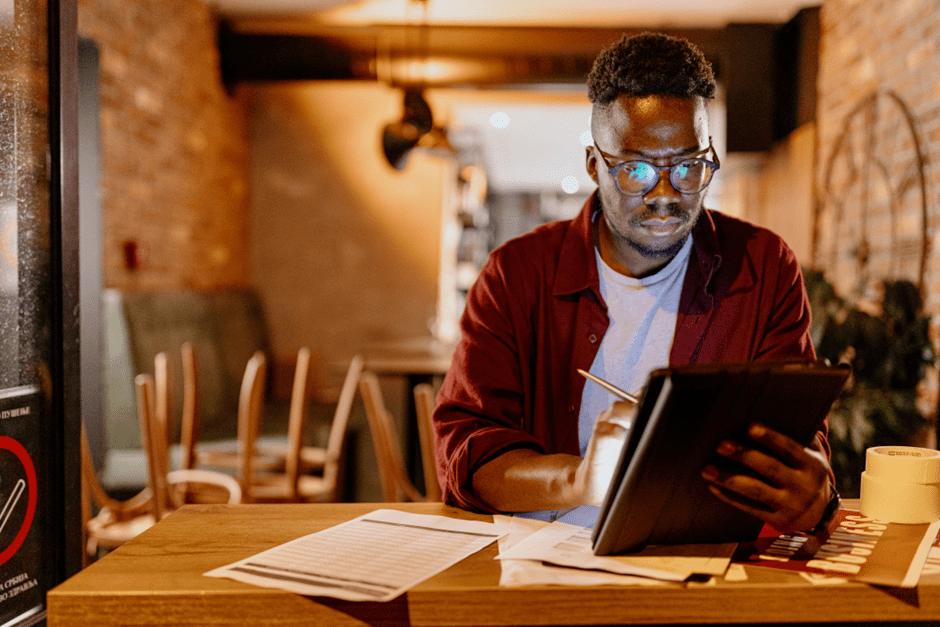 A cafe/bar owner, stood at a table looking at an electronic iPad, with papers scattered on the table