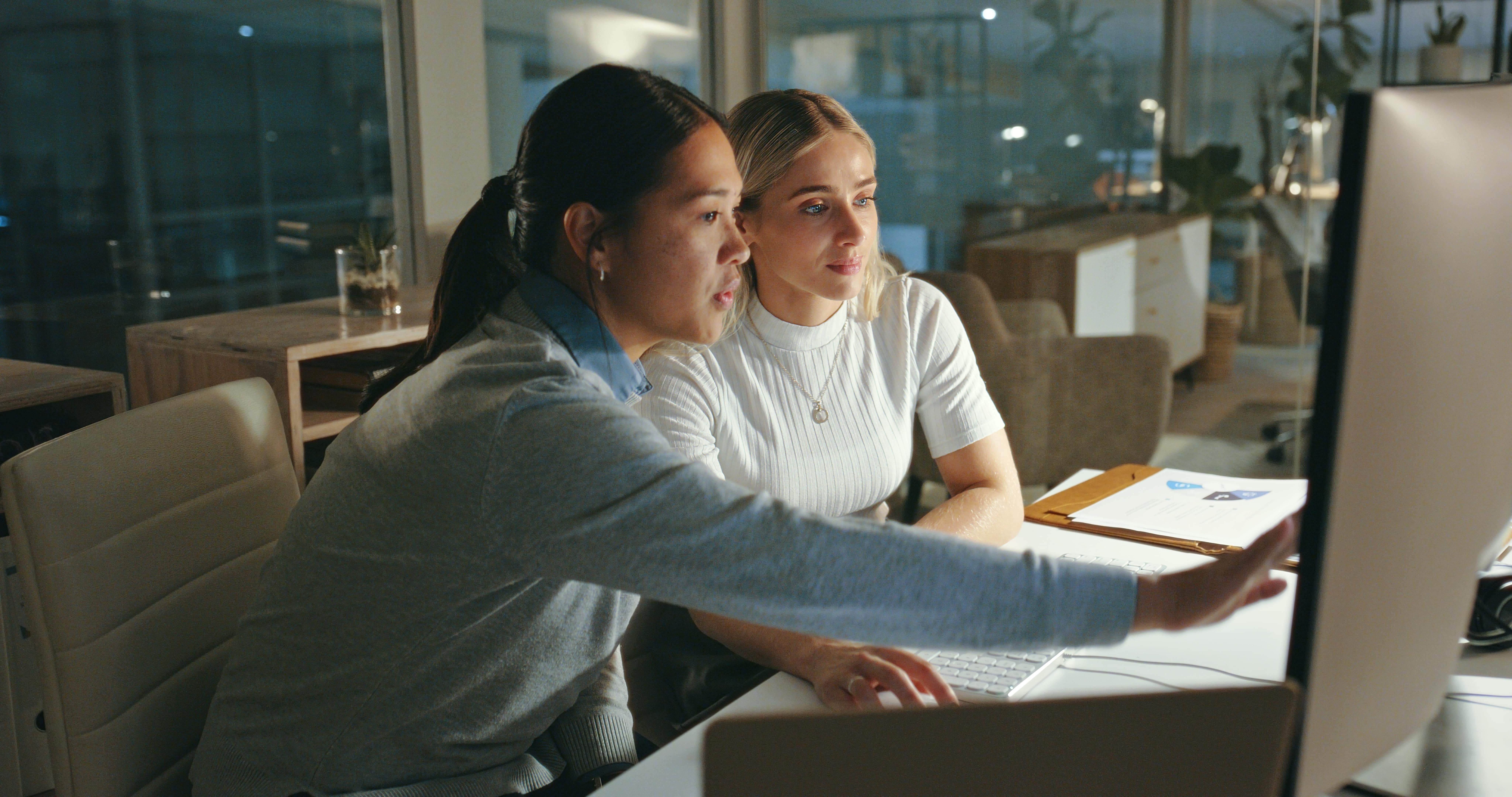 Two people sat in an office infront of a computer, with one person pointing at the computer screen