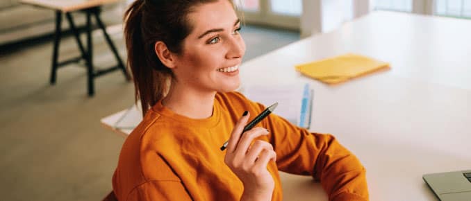 Smiling woman in an orange sweater sitting at a desk, holding a pen.