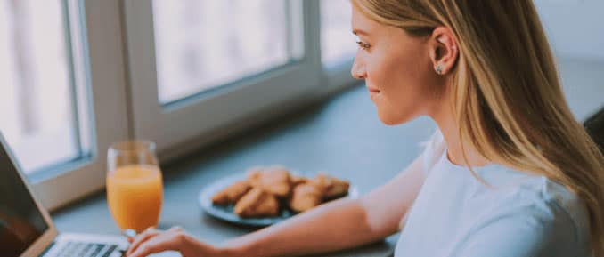 Young woman working on a laptop near a window, with a glass of orange juice and a plate of pastries in the background.