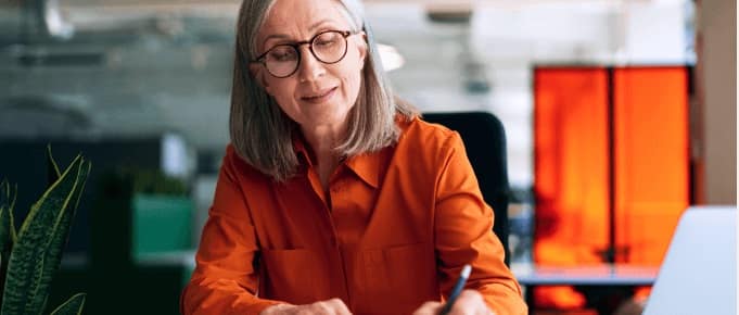 Woman wearing glasses and an orange shirt, writing at a desk in a modern office setting.