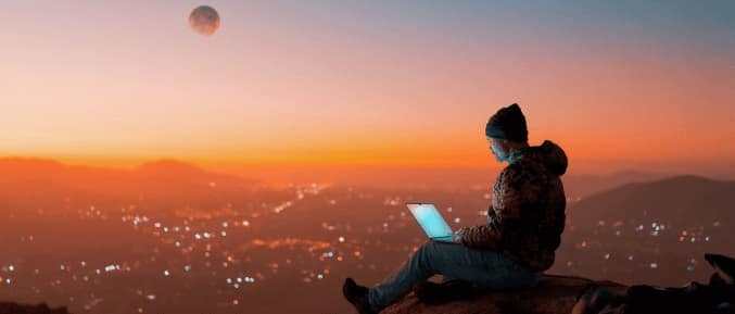 Person sitting on a mountain at sunset, using a laptop, with a cityscape the background.