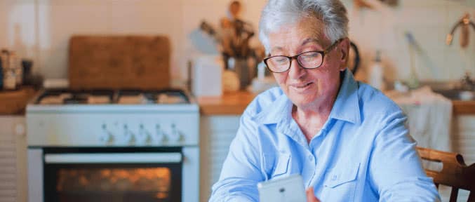 Elderly woman with glasses sitting in a kitchen, smiling while looking at her smartphone.