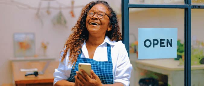 Self-employed small business owner smiling outside her shop with an open sign, holding a smartphone