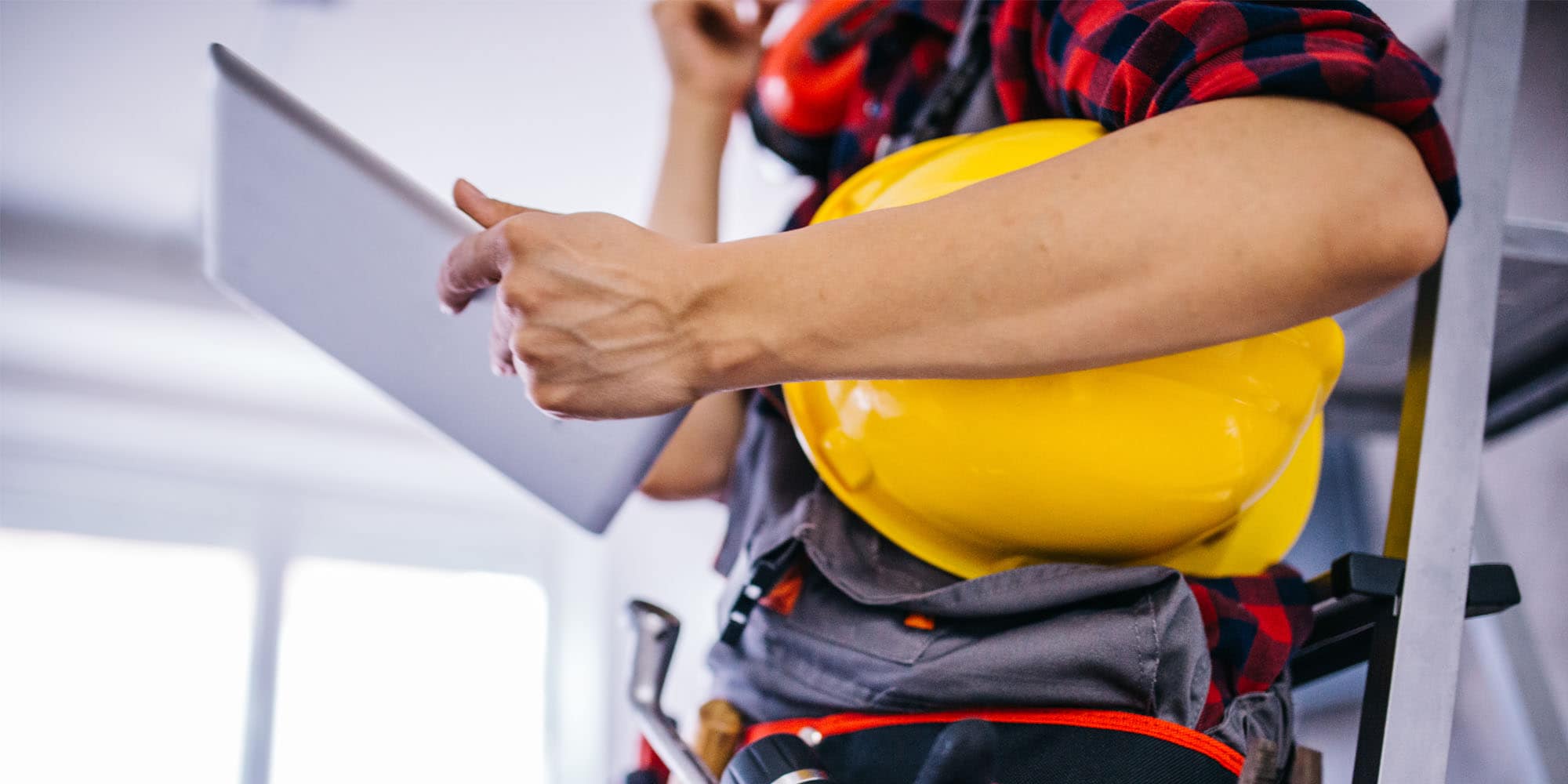 Construction worker holding a yellow hard hat and reviewing a tablet at a job site