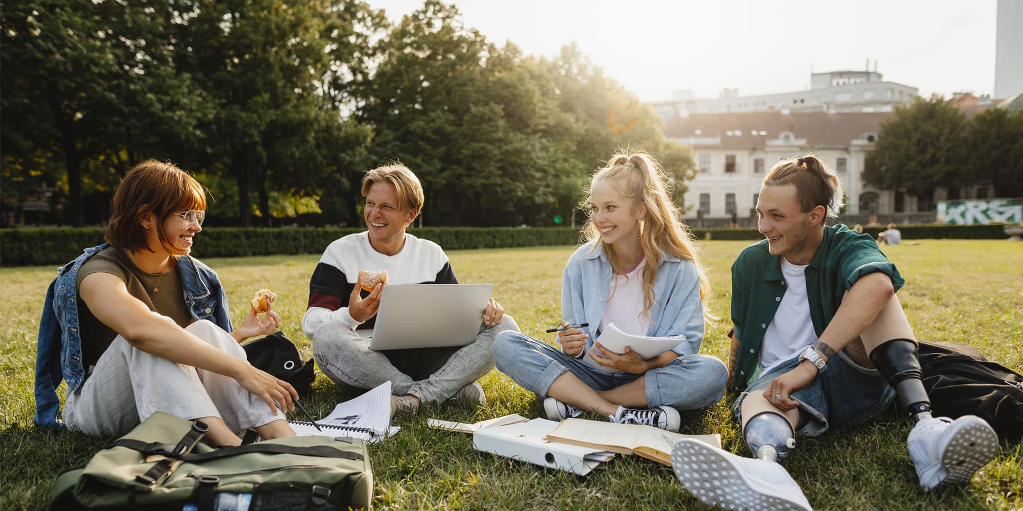 Group of people sitting on grass in a park, with notebooks and a laptop spread out in front of them