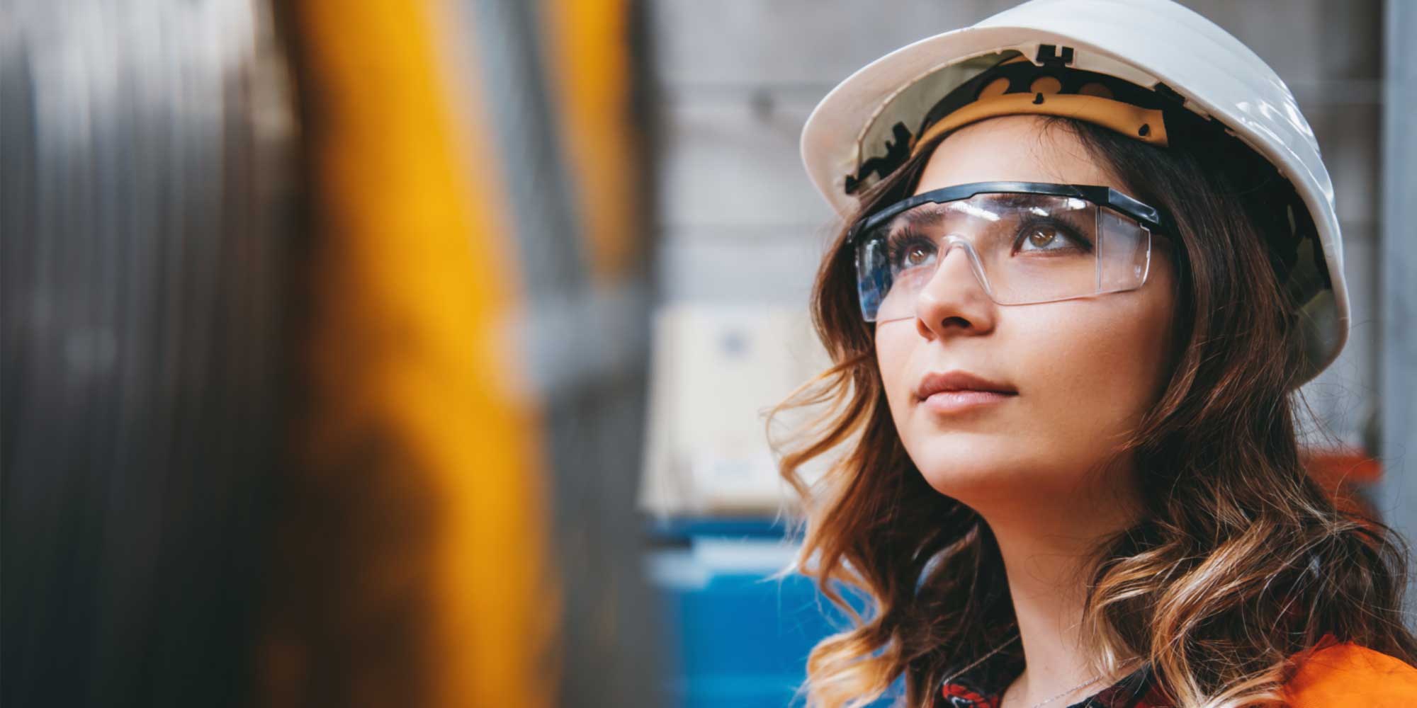 Person wearing a hard hat and safety glasses looking upward inside an industrial facility