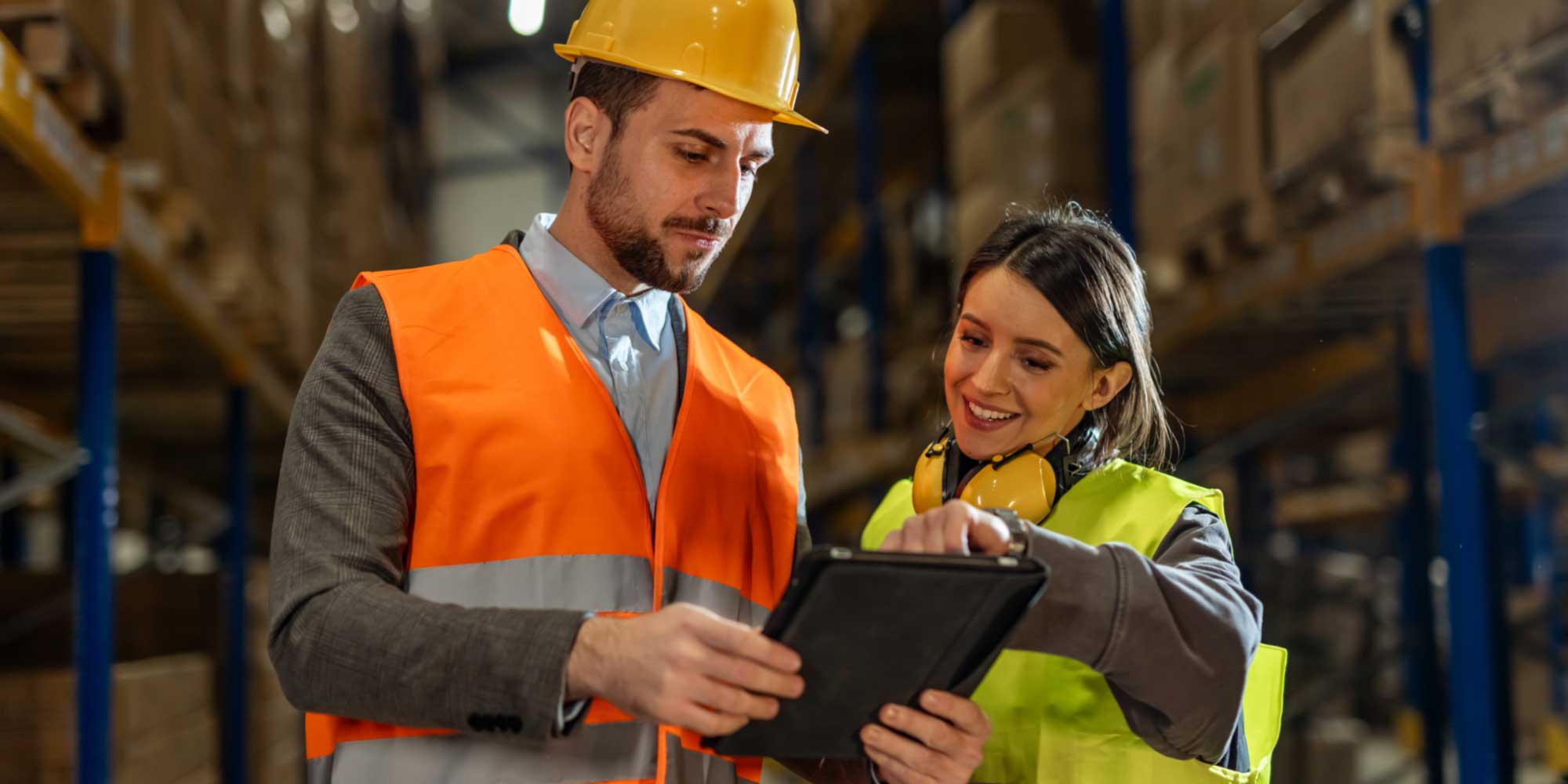 Two people wearing safety gear reviewing information on a tablet in a warehouse