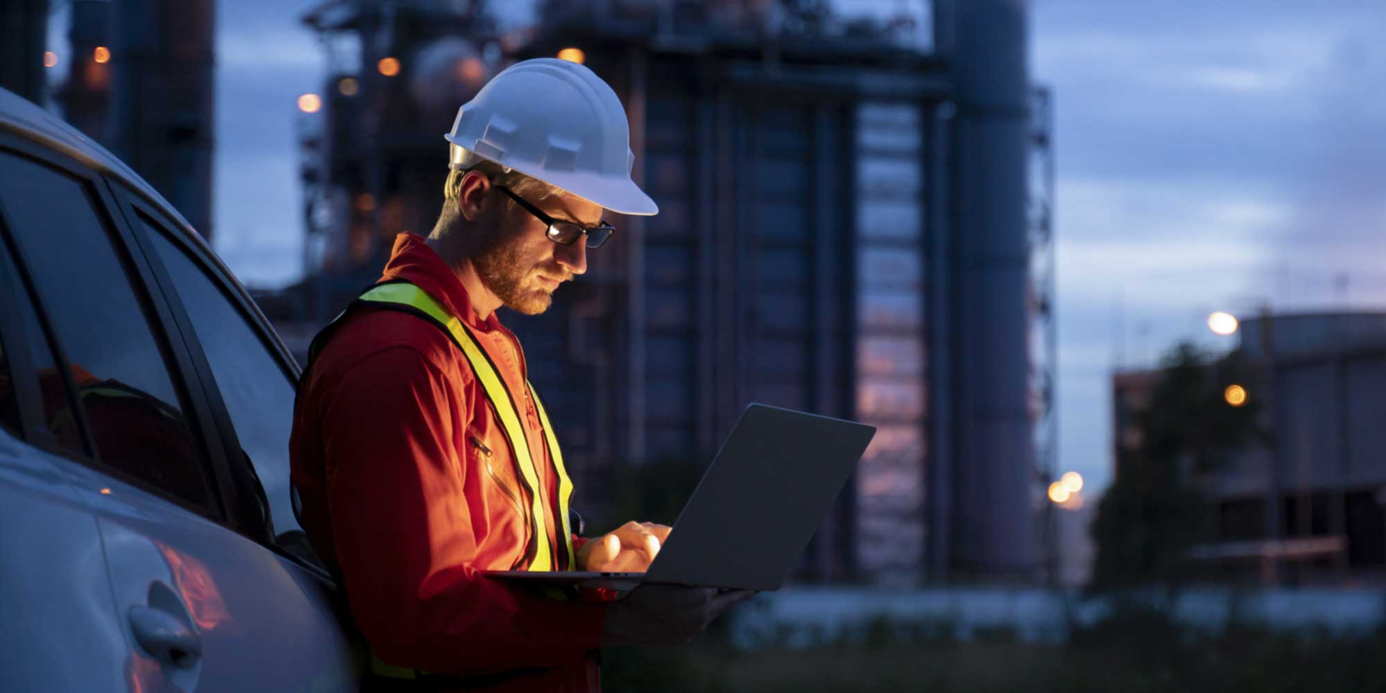 Person wearing a hard hat and high‑visibility vest using a laptop near an industrial facility at dusk