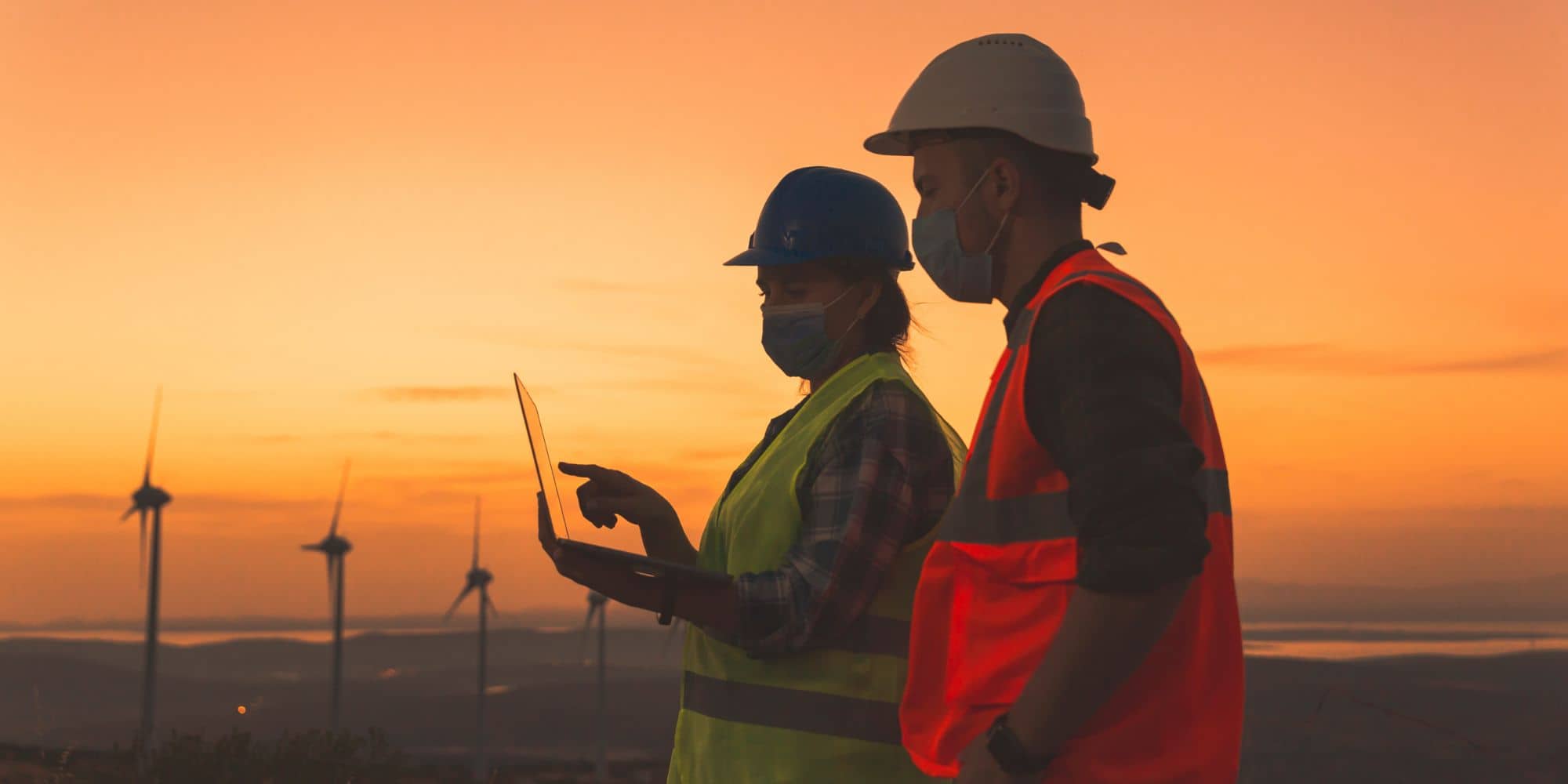 Two workers in safety gear using a laptop at a wind farm during sunset.