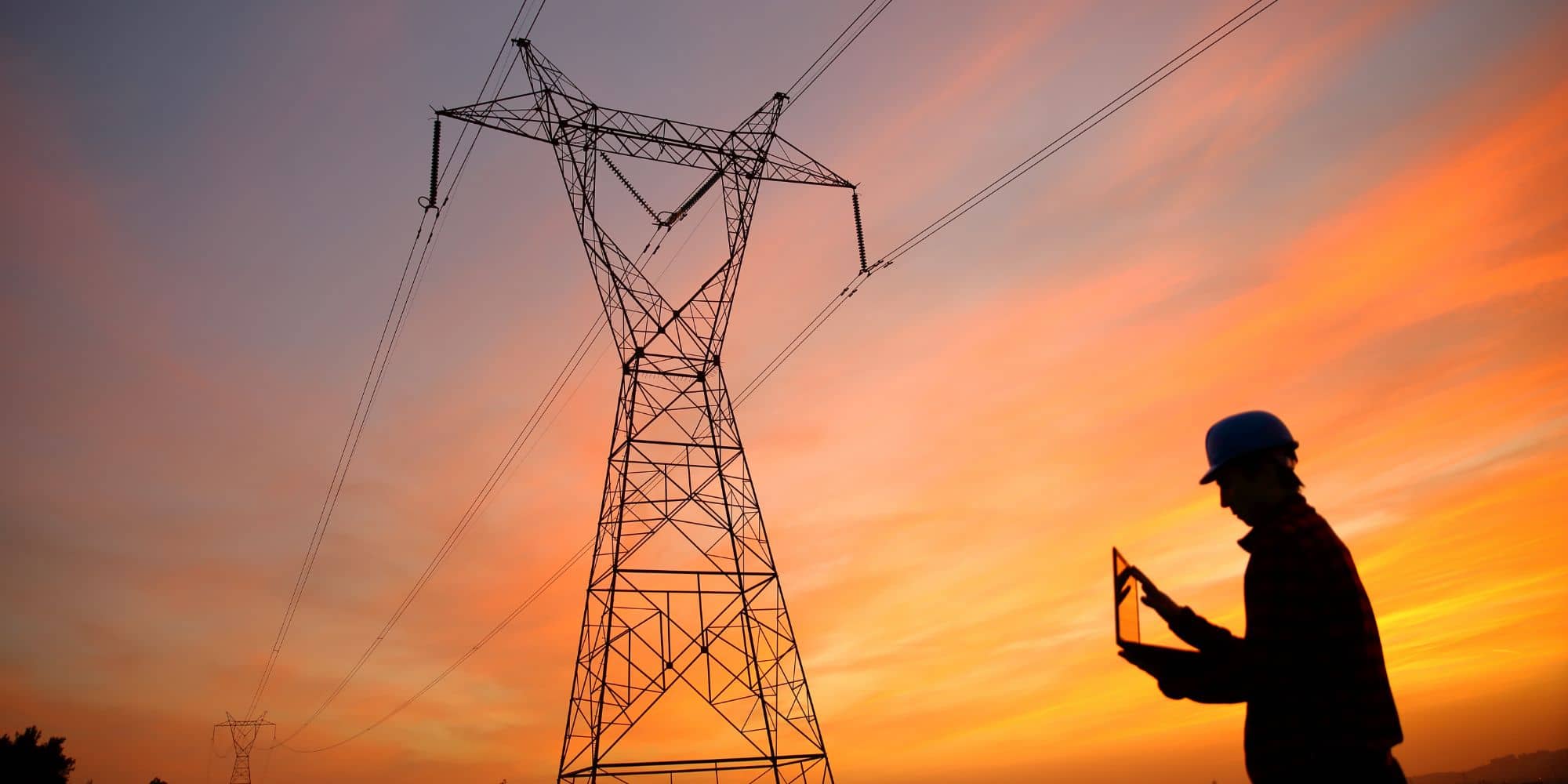 A silhouette of a person with a hard hat holding a tablet near an electrical transmission tower during sunset.