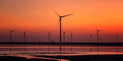 A serene landscape at sunset featuring numerous wind turbines. The sky is painted in shades of orange and red, reflecting on the calm water below. The silhouettes of the wind turbines stand tall against the colorful backdrop, creating a peaceful and picturesque scene that highlights renewable energy sources.