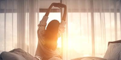 Woman waking up and stretching in bed with sunrise in the background